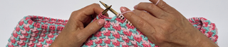 Close-up of a person's hands knitting with wooden needles and a pink, teal, and white textured yarn.